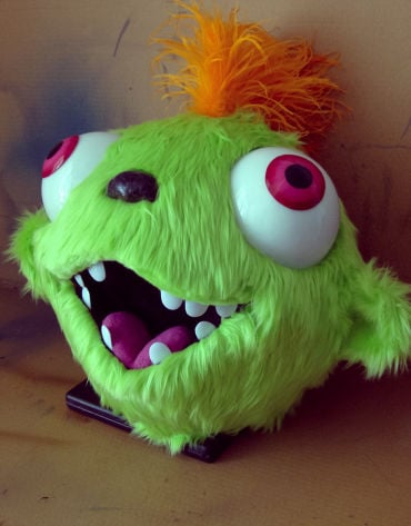 Head of a mascot costume with green fur, bulgy magenta eyes and orange hur