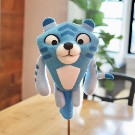 Front view of a custom blue tiger puppet with big eyes, a white belly and muzzle, blue stripes, and small rounded ears, resting on a wooden stand.