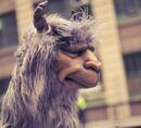 Side-profile photo of a gray shaggy llama puppet with twisted dark horns, closed eyes, a soft sculpted muzzle. In the background: Industry City buildings in Brooklyn, New York.