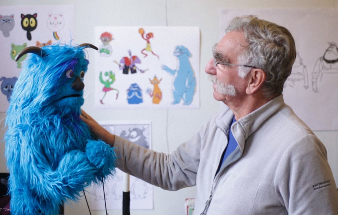 Puppet maker at his studio, looking at one of his creations: A blue puppet with two horns and surprised expression.