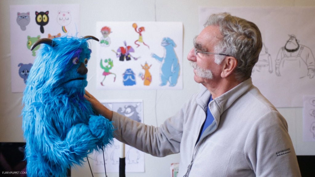 Puppet maker at his studio, looking at one of his creations: A blue puppet with two horns and surprised expression.