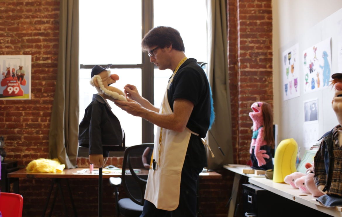 An artist at works in a New York puppet studio. Assorted hand puppets in the background.